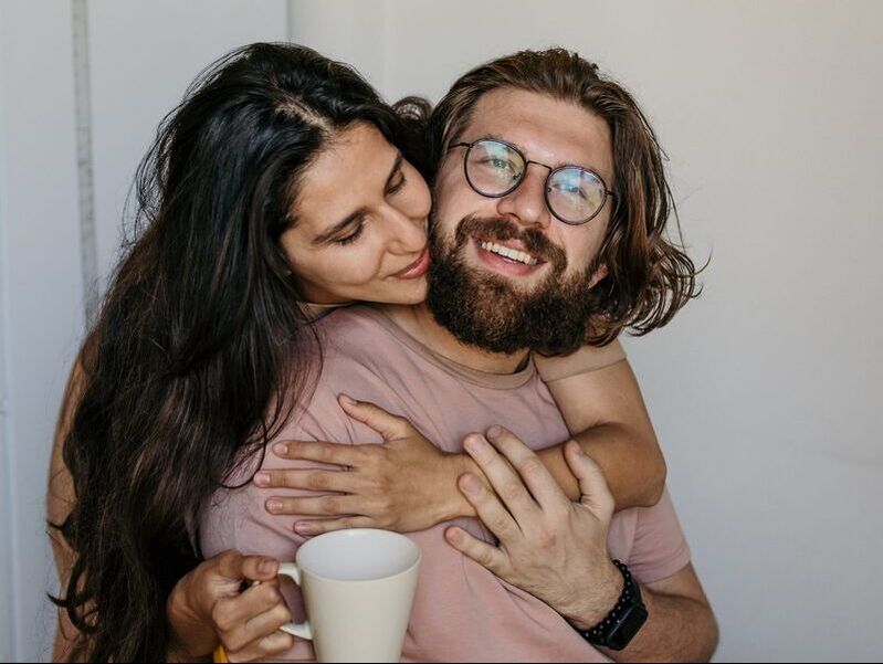 A man and woman enjoy a cozy hug, both holding a cup of coffee together.